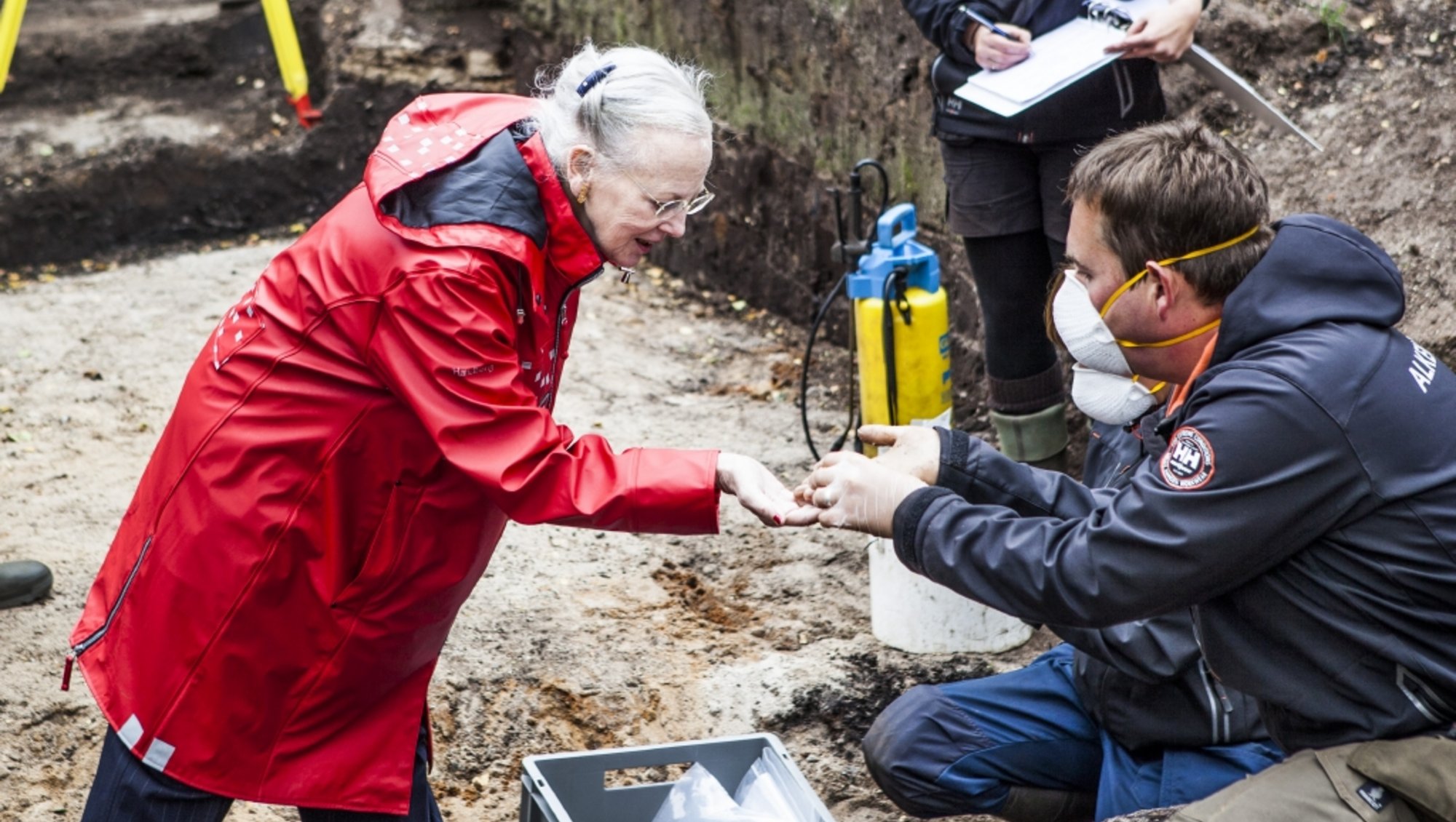 Dronning Margrethe, iført en rød regnfakke, står i en udgravning og en person i blåt regntøj og maske, viser hende noget, der er fundet i udgravningen. 